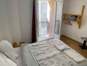 A bedroom with a bed, a wooden floor, a window with a metal gate, and a wooden shelf with clothes. - Hostel Le Diablotin (Saint-Jean-de-Fos)