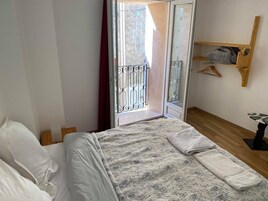 Un dormitorio con cama, suelo de madera, una ventana con celosía metálica y estante de madera con ropa.