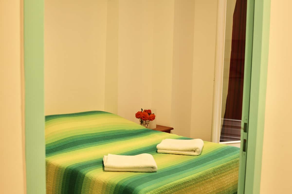A neatly made bed with a striped bedspread and two folded white towels. A small table with a vase of red flowers is visible in the corner.