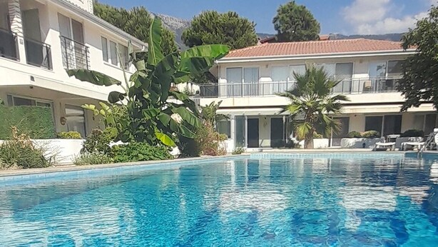 A clear blue pool with tiled edges, surrounded by lush greenery and a two-story building with balconies. - Oludeniz Evleri (Fethiye)