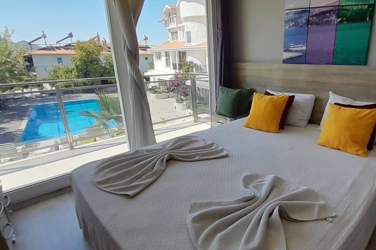 A bedroom with a bed, pillows, a view of a swimming pool, and a balcony.