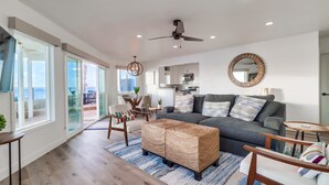 A modern living room with a grey sofa, wicker ottoman, and a dining area with chairs and a table.