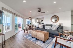 A modern living room with a grey sofa, wicker ottoman, and a dining area with chairs and a table. - Oceanfront new listing with Spectacular Views (San Diego)