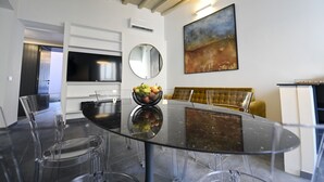 A modern dining area with a glass table, clear chairs, a bowl of fruit, a television, a mirror, and a painting on the wall. - Sistina36 Spanish Steps Apartment (Roma)