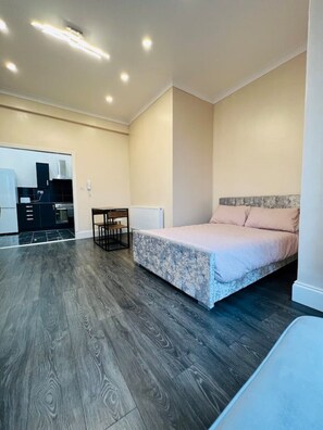 Un dormitorio con una cama, una mesita y una kitchenette al fondo.
