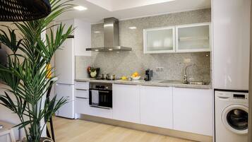 A modern kitchen with white cabinets, a stainless steel range hood, a built-in oven, a dishwasher, and a washing machine.