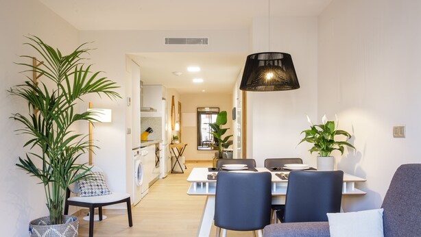 En moderne stue med en grå sofa, et spisebord med stole og et køkken i baggrunden.