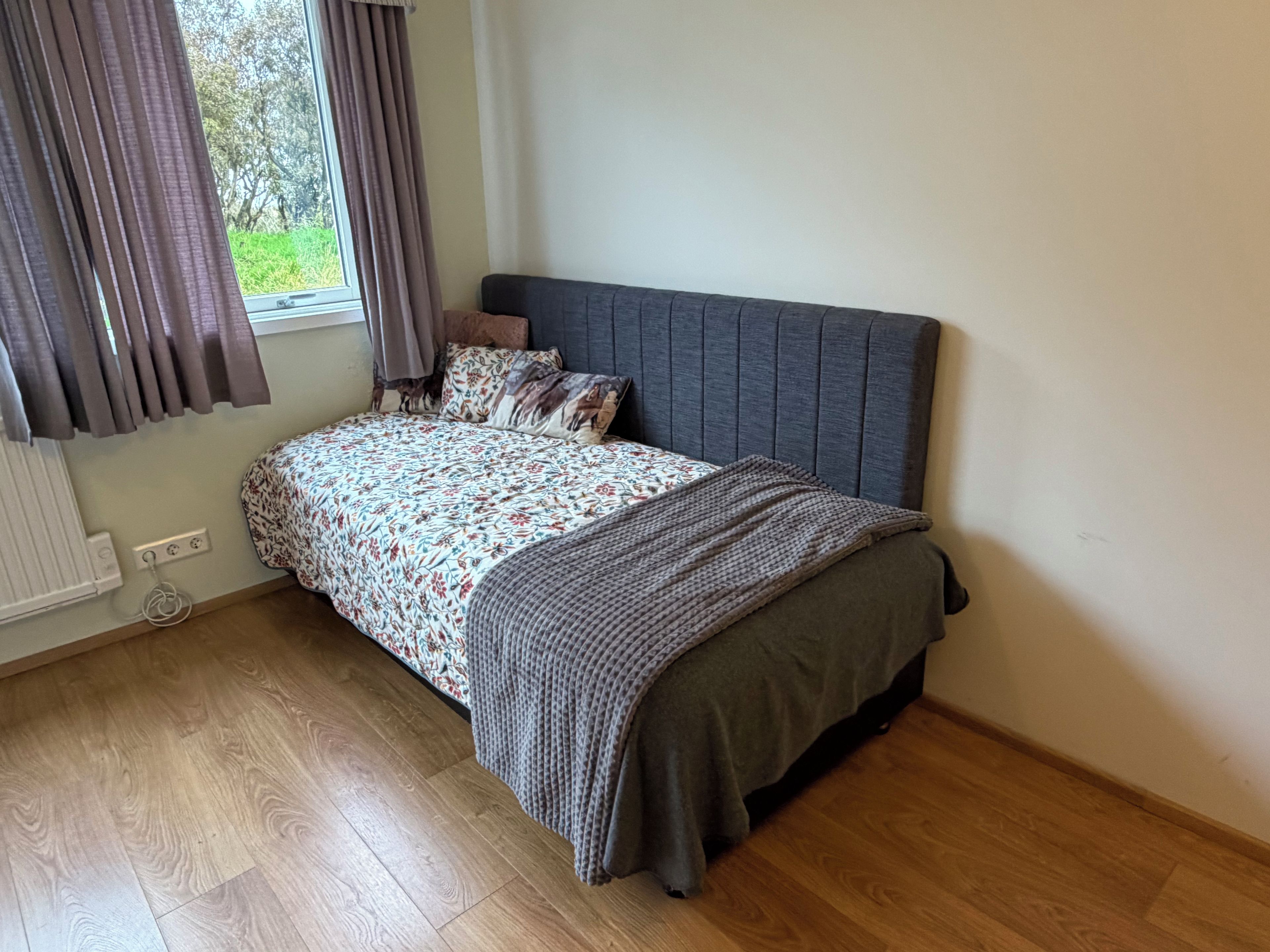 A bedroom with a bed, a window with curtains, a radiator, and wooden flooring.