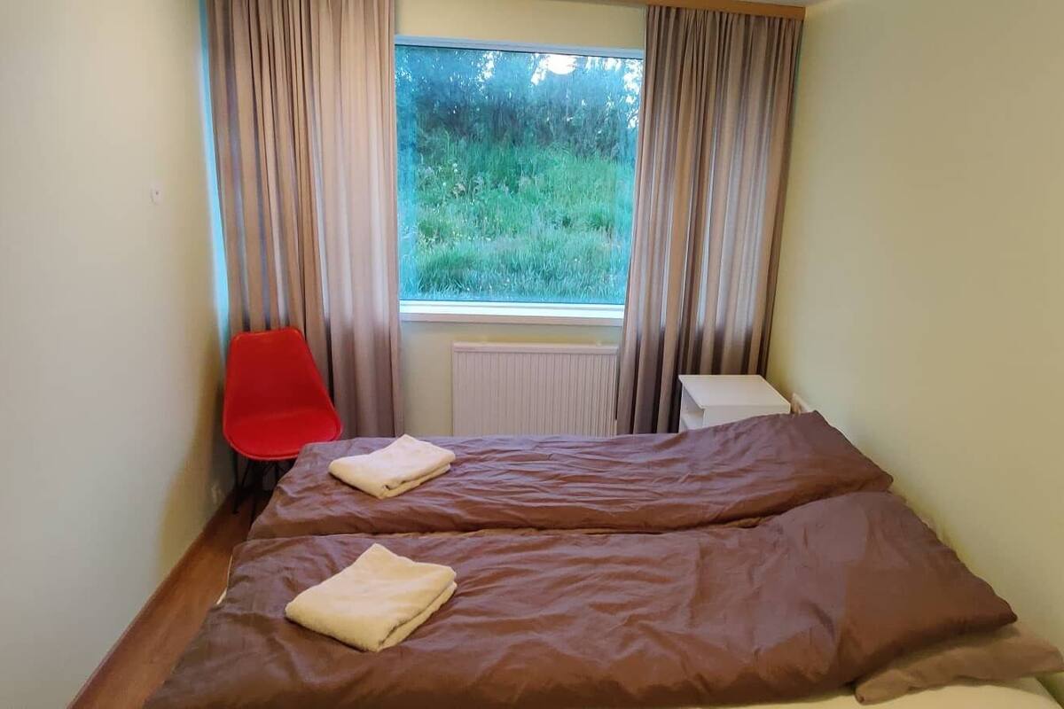 Une chambre avec deux lits, un fauteuil rouge et une fenêtre avec des rideaux.