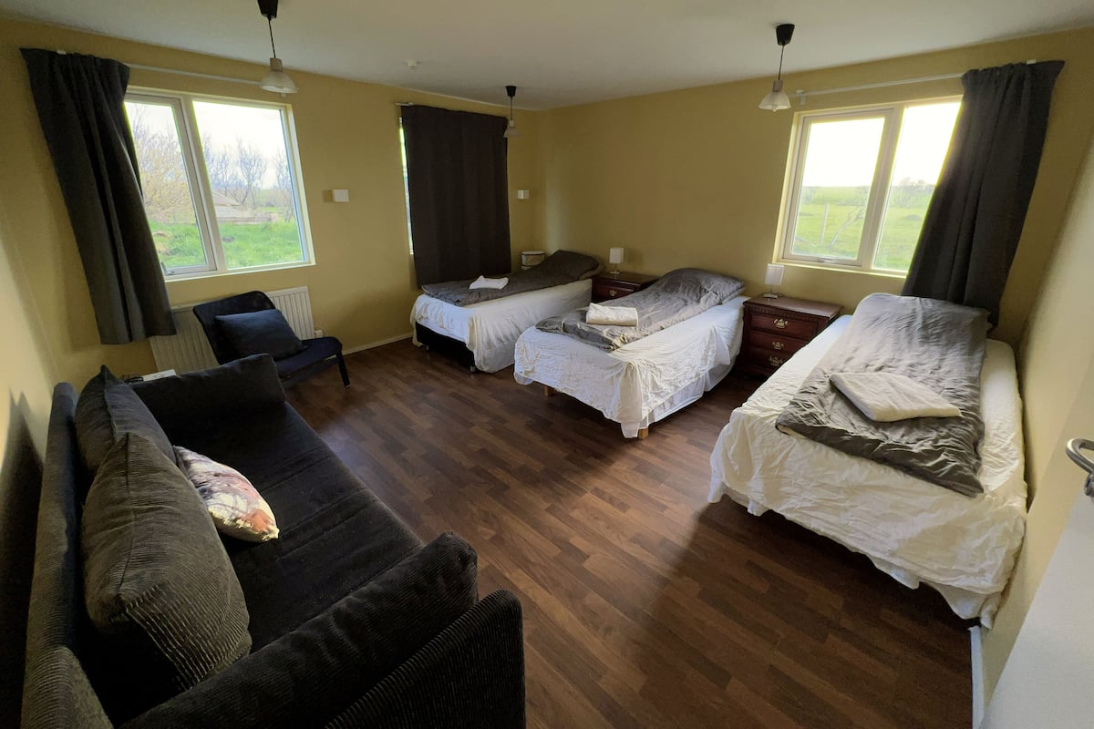 Une chambre avec deux lits simples, un canapé et un fauteuil. Elle est équipée de fenêtres avec des rideaux, d’un radiateur et d’un ventilateur de plafond.