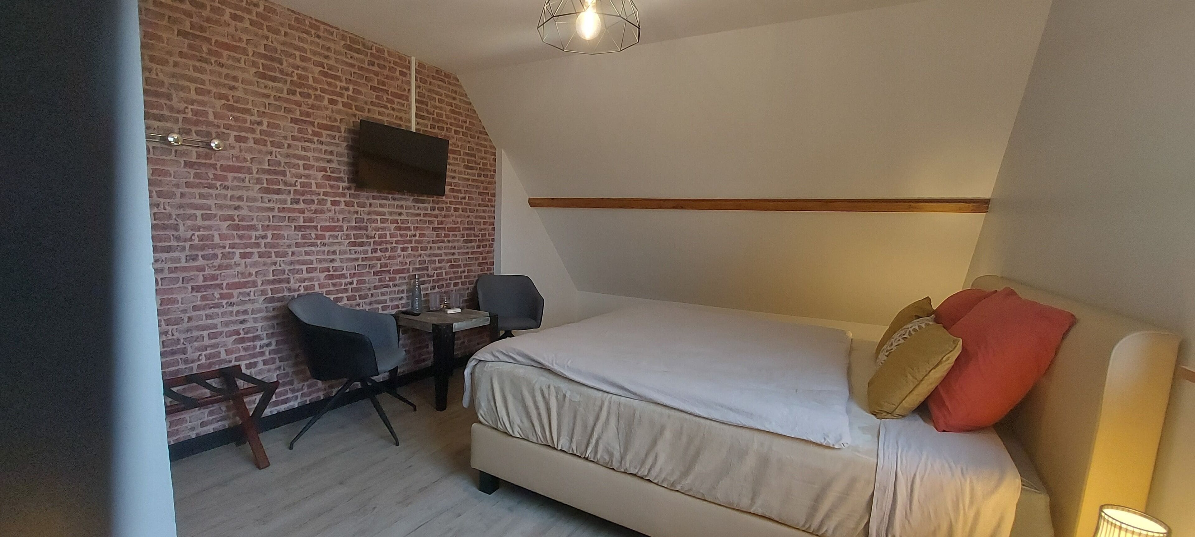 A bedroom with a brick wall, a bed with pillows, a desk with a chair, and a mounted TV.