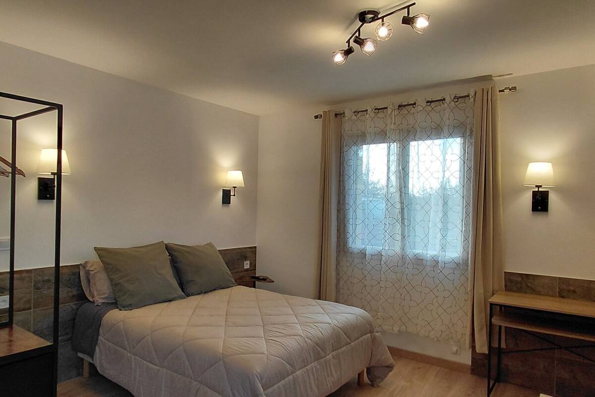 Une chambre à coucher comprenant un lit, une table de chevet, un miroir, une armoire, un téléviseur fixé au mur et une fenêtre avec des rideaux.