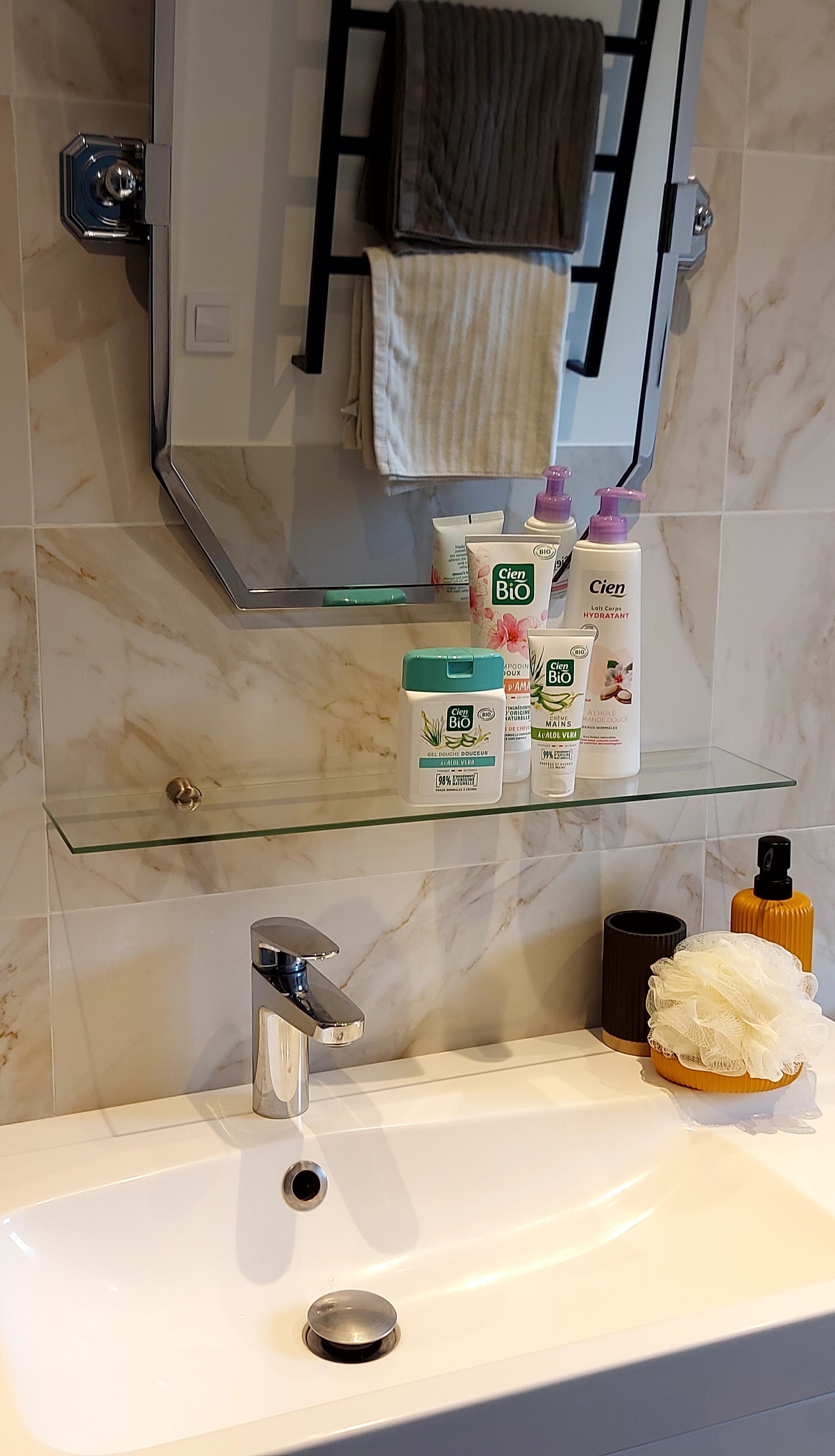 Un baño con un estante de vidrio que sostiene artículos de tocador, un lavamanos con un grifo moderno y un perchero sobre el espejo.