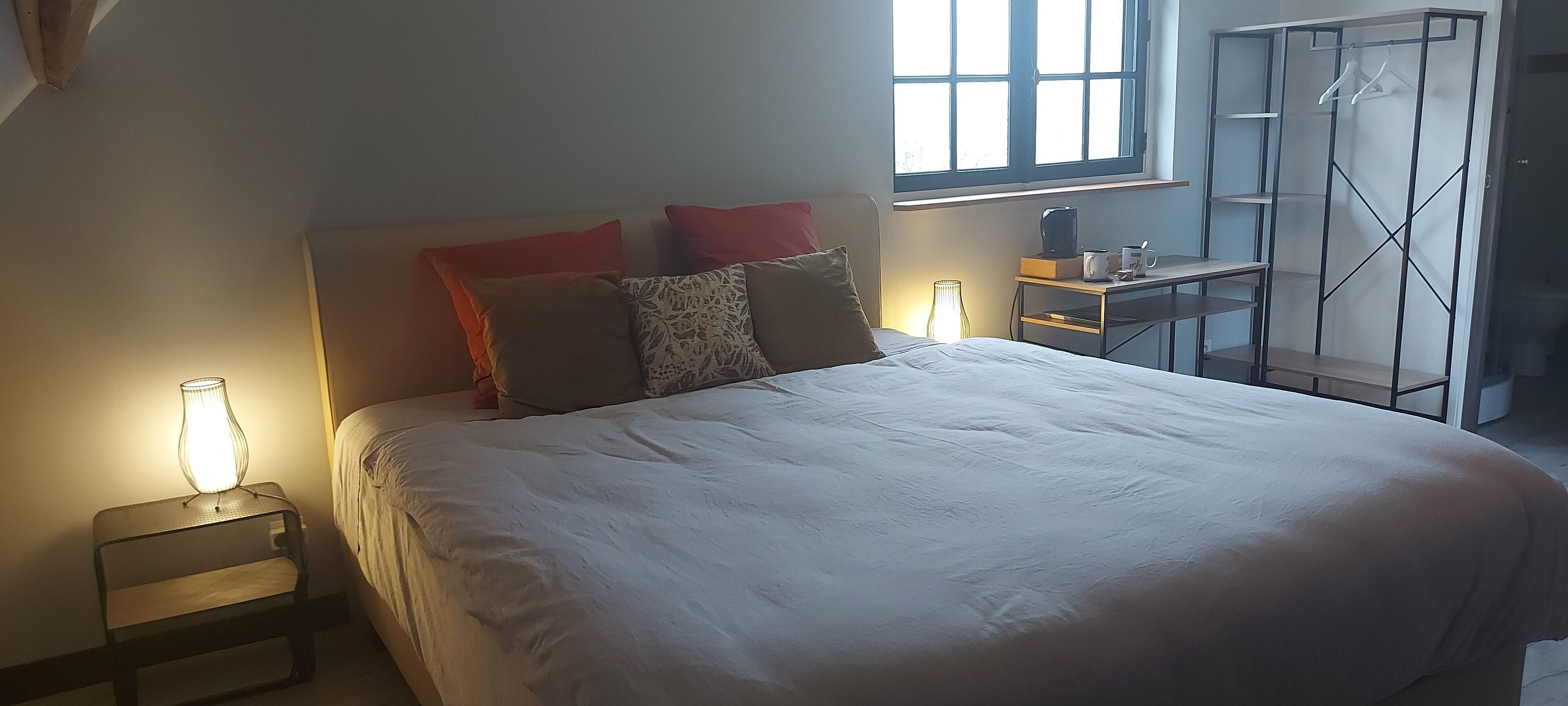 A neatly made bed with pillows, a bedside table with a lamp, and a window with natural light.
