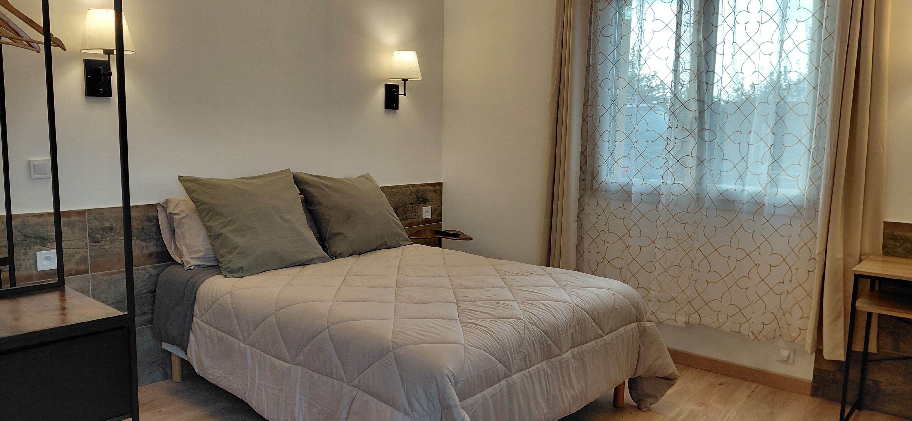 A neatly made bed with a quilted cover, a bedside table with a lamp, and a window with patterned curtains.