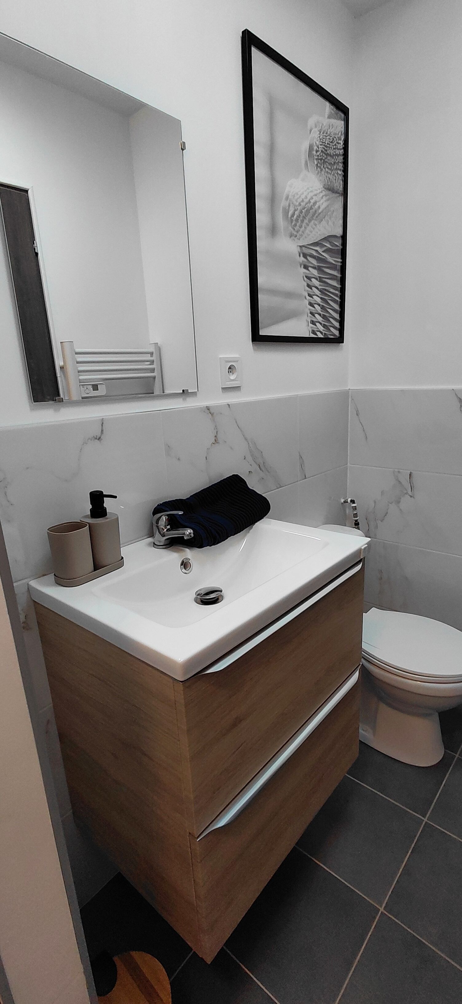 Un baño moderno con un lavamanos blanco, tocador de madera, una toalla negra, un inodoro blanco y un cuadro en la pared.