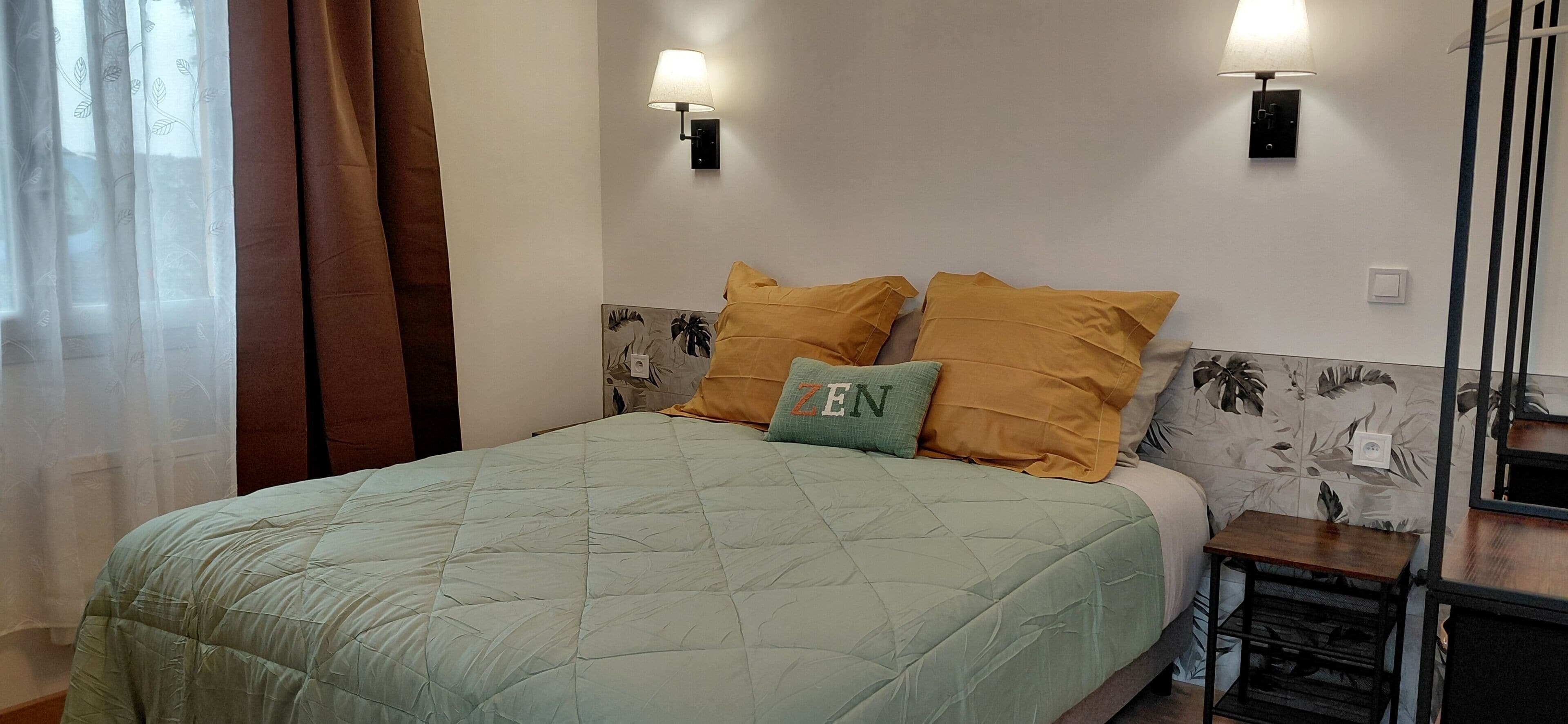 A bed with a green quilt, two wall-mounted lamps, and a small table beside it.
