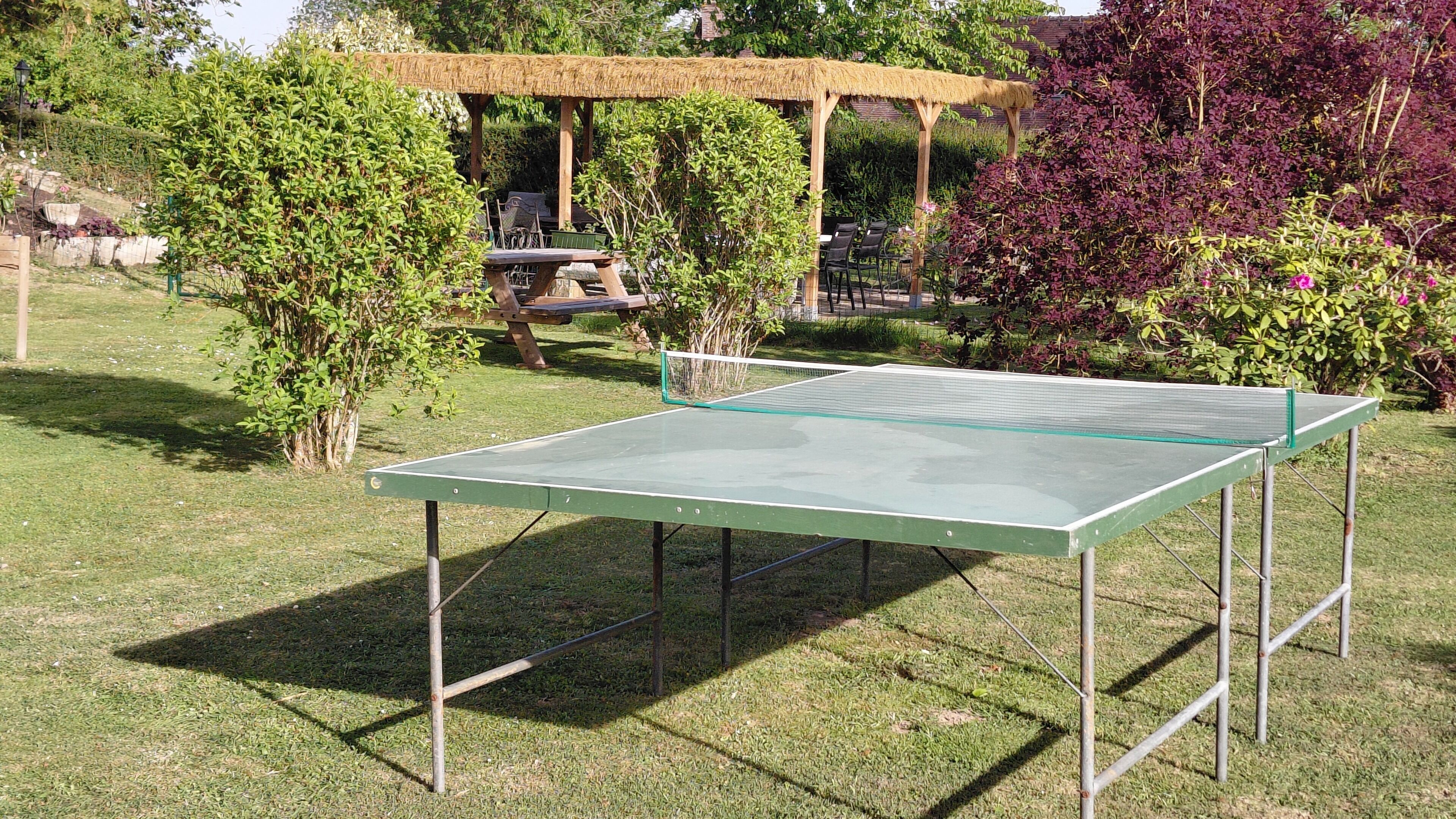 Mesa de pingue-pongue ao ar livre num jardim, com uma pérgula e zona de estar ao fundo.