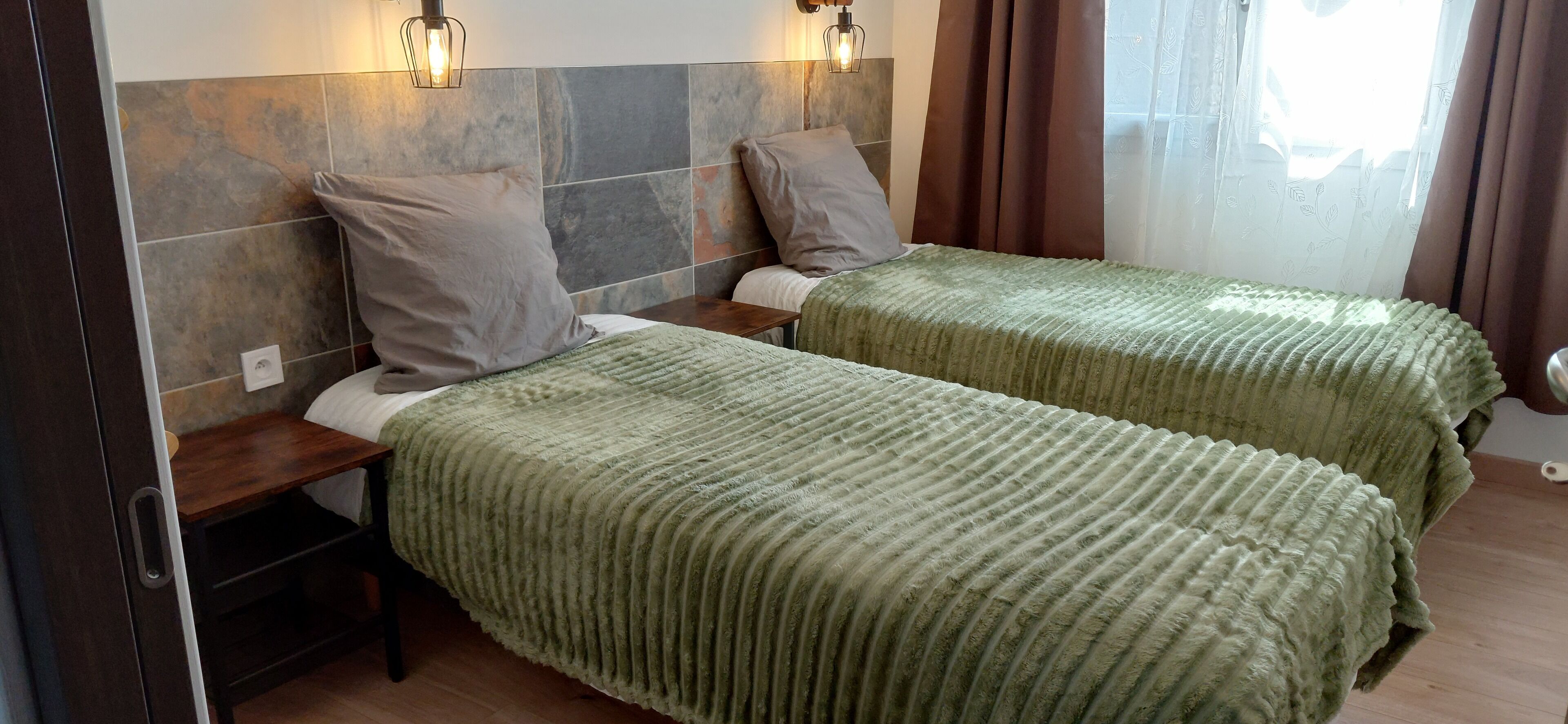 Hotelkamer met twee bedden, elk met een groene sprei en kussens, een houten nachtkastje en een wandlamp.