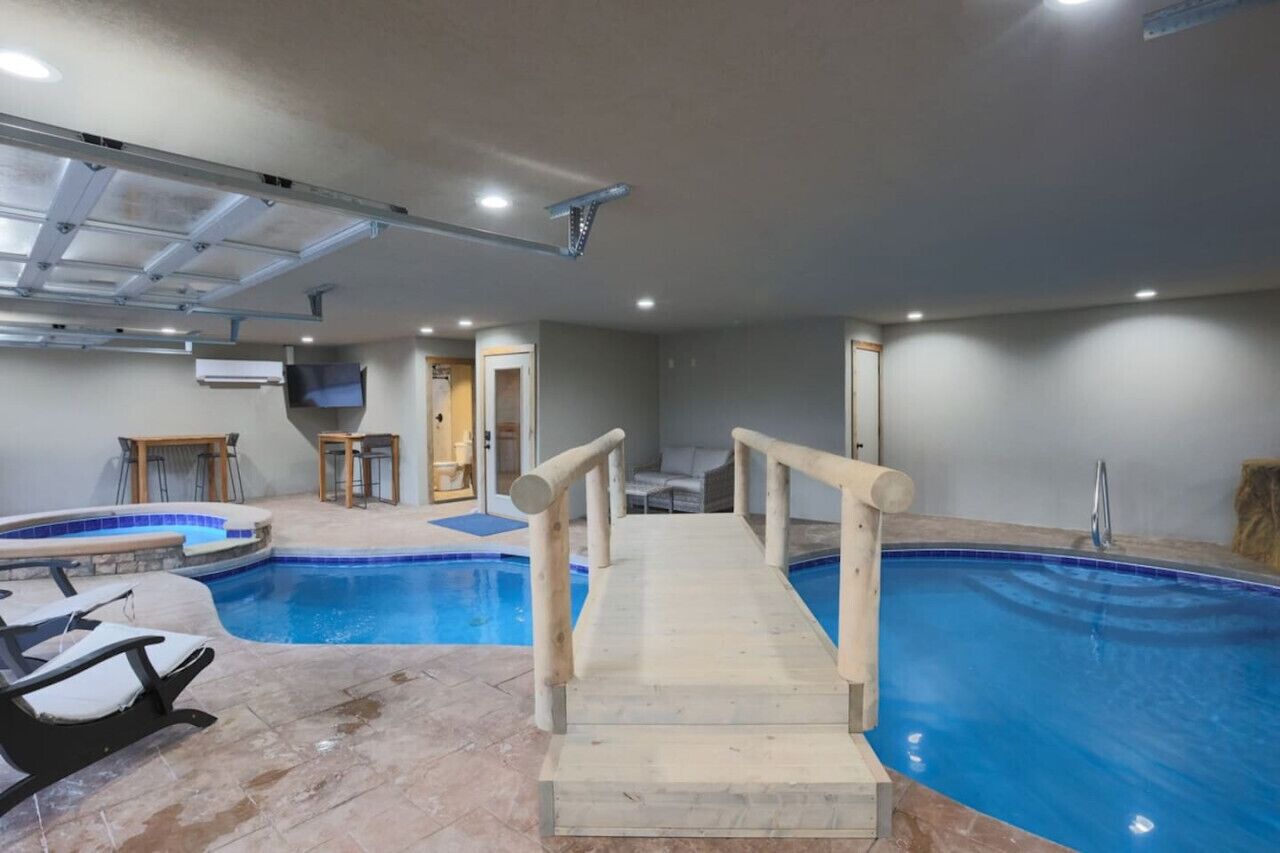 An indoor swimming pool with a wooden deck and railings, surrounded by a tiled floor and a ceiling with recessed lighting.