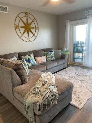 Un salon avec un canapé d’angle gris, une décoration murale représentant une boussole, une porte vitrée donnant sur l’extérieur et une chaise Adirondack verte visible à travers la porte.