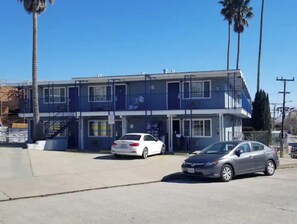 Two-story building with a flat roof and balcony, surrounded by palm trees and lush vegetation.