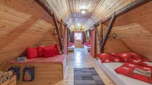 Une pièce mansardée chaleureuse aux murs en bois et aux plafonds inclinés, comprenant deux lits avec une literie rouge, un petit coin salon et une fenêtre avec des rideaux.