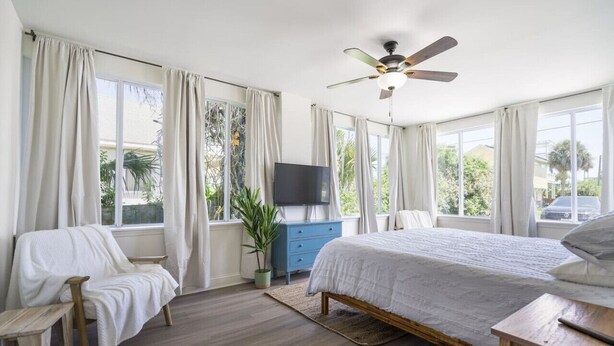 Un dormitorio con cama, televisor, ventilador de techo y ventanas amplias.