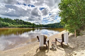 A lakeside view with two chairs and a table, a bottle, and a bottle of wine on the sand.