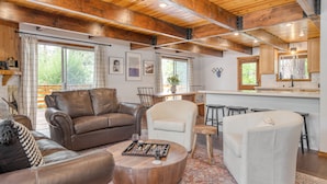Una sala de estar acogedora con un sofá de cuero marrón, un sillón blanco y una mesa de centro de madera. Hay una zona de cocina con una barra y taburetes.