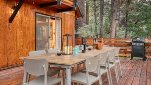 Una terraza de madera con mesa y sillas, una parrilla y vista a árboles.