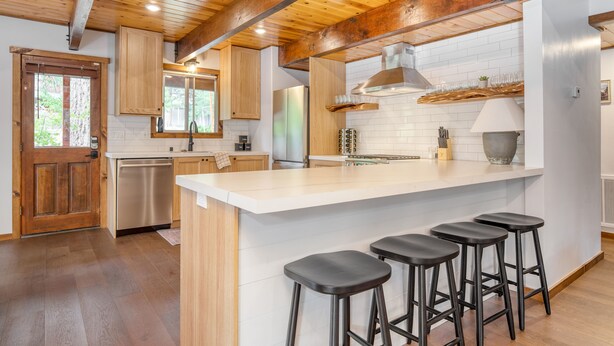 Una cocina moderna con vigas de madera, una isla grande con taburetes de bar, electrodomésticos de acero inoxidable y un salpicadero de azulejos blancos.