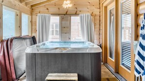 Interior de una cabaña de madera con jacuzzi, ventanas amplias y cortinas.