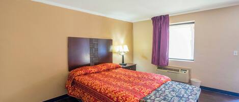 A hotel room with a bed, a nightstand, a lamp, a window with curtains, and a bench.