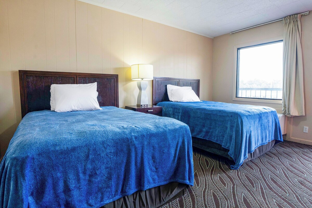 A hotel room with two beds, a nightstand, a lamp, and a window with curtains.