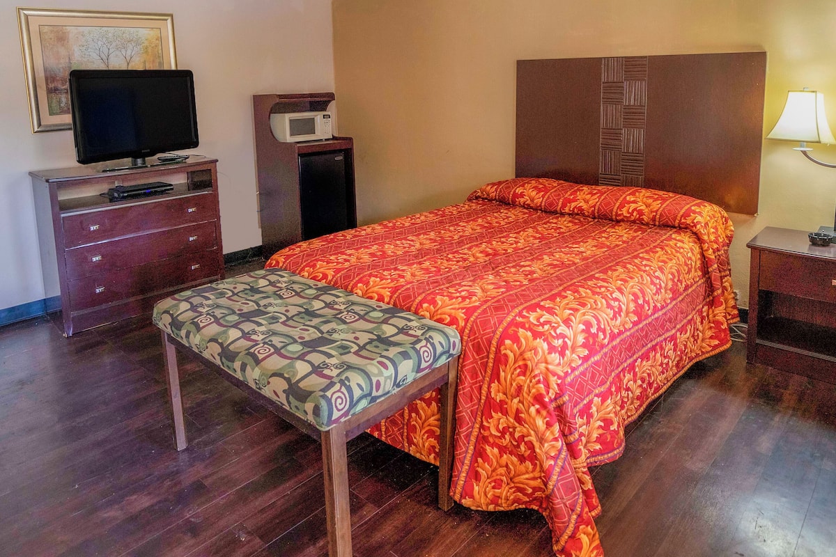 Habitación de hotel con cama, televisión en una cómoda, microondas y mesita de noche con lámpara.