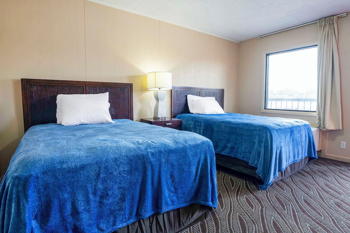 Una habitación de hotel con dos camas, una mesita de noche, una lámpara y un ventanal con cortinas.