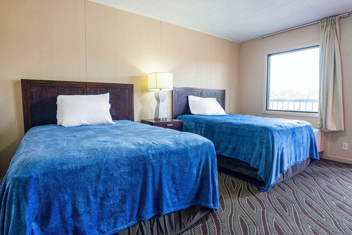 A hotel room with two beds, a nightstand, a lamp, and a window with curtains.