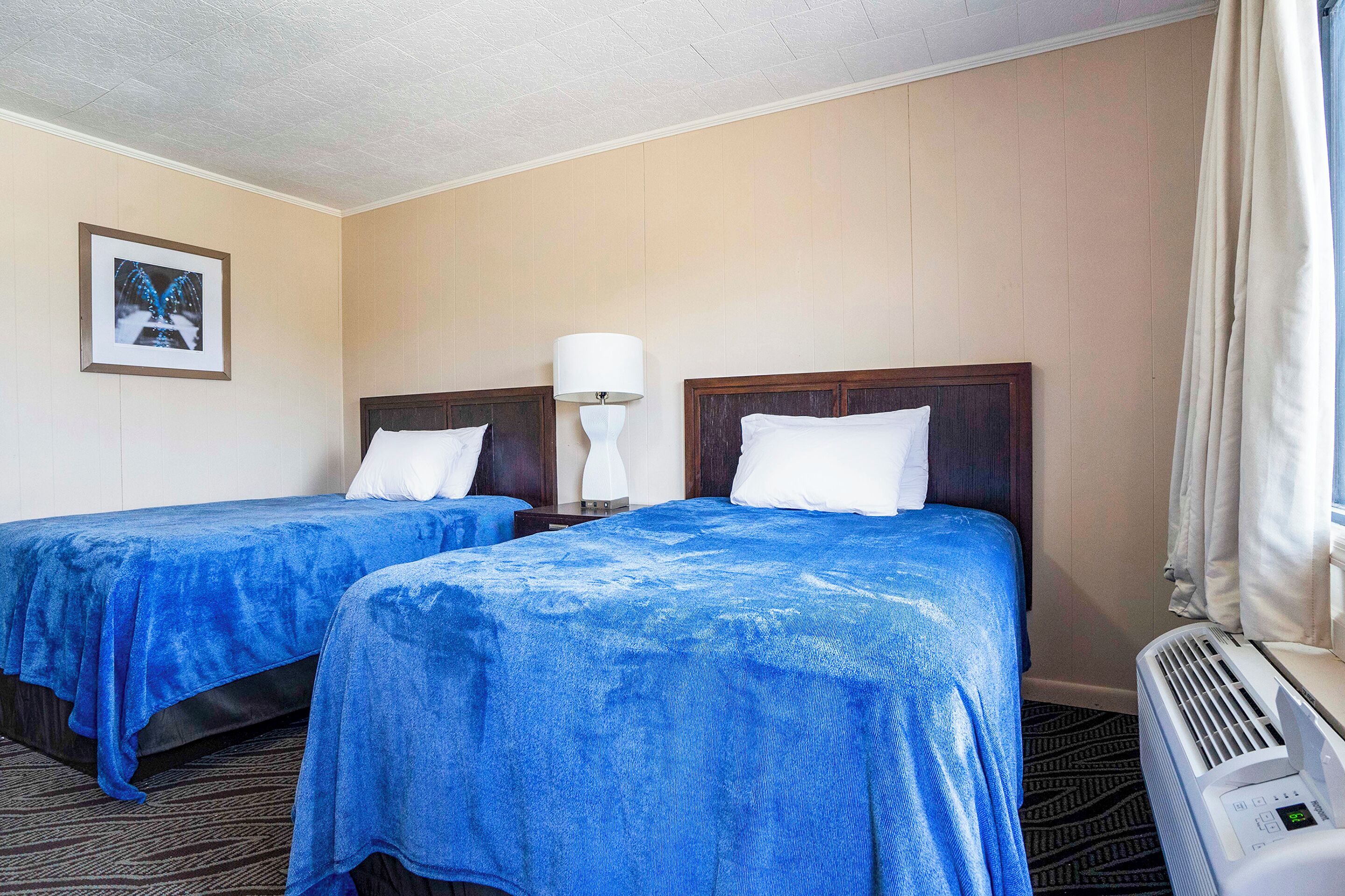 Una camera d'albergo con due letti, un comodino con lampada, un quadro appeso al muro e una finestra con tende.