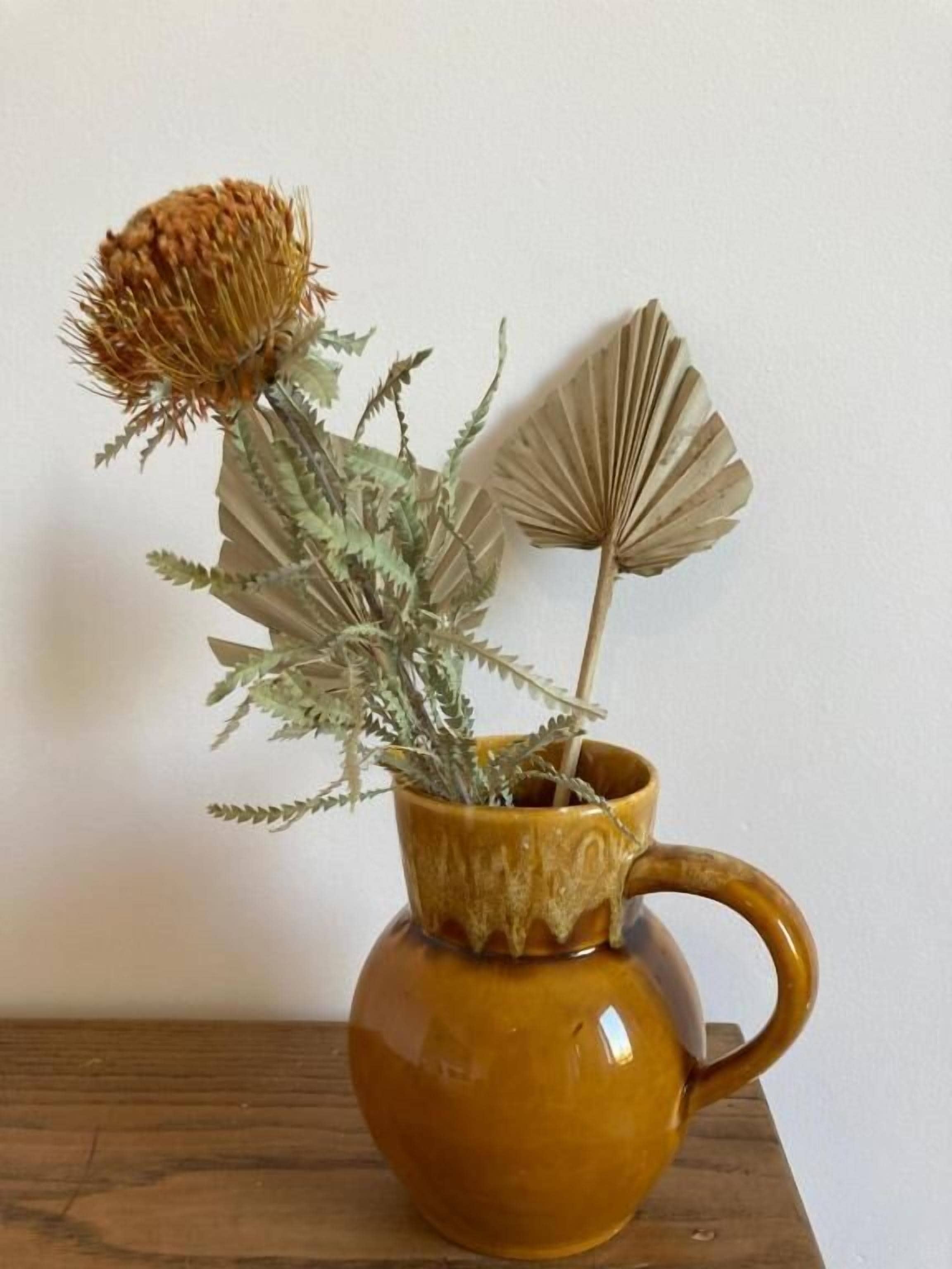 A brown ceramic mug with a handle, containing dried plants and a fan-shaped paper decoration.