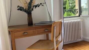 Ein Holztisch mit einem gewebten Stuhl, eine Vase mit Trockenblumen und Blick ins Grüne durch ein Fenster.
