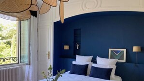 A bedroom with a large bed, white bedding, and dark blue walls. There is a window with a view of greenery, a radiator, and a vase with flowers on the bed. - La Maison 19 (Niort)