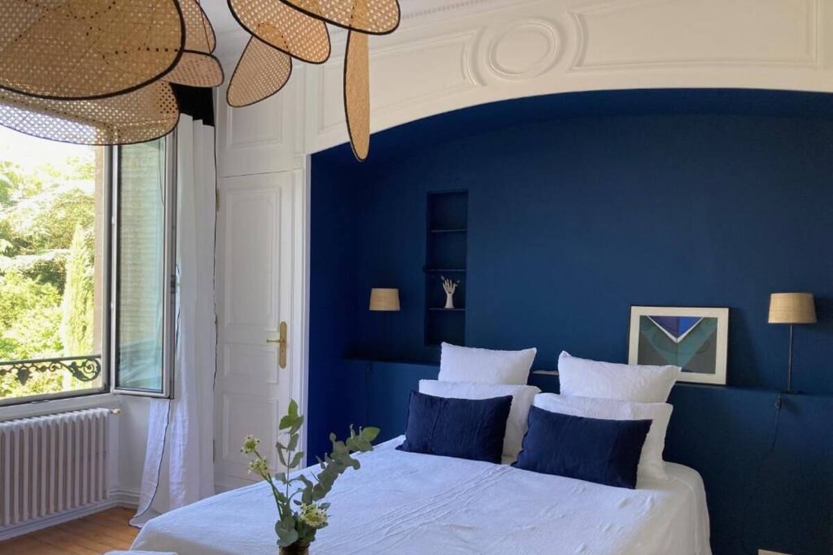 Un dormitorio con una cama grande, ropa de cama blanca y paredes azul oscuro. Hay una ventana con vistas a zonas verdes, un radiador y un jarrón con flores sobre la cama.