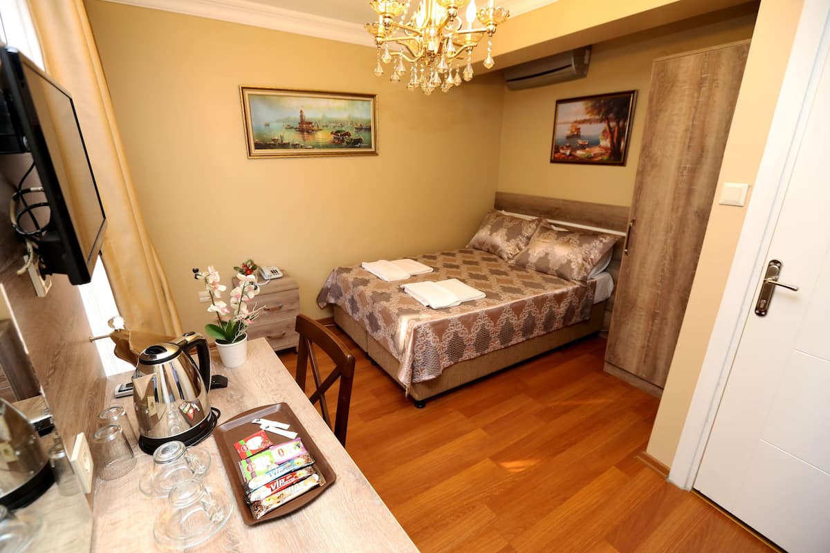 A bedroom with a bed, a desk with a kettle, a chair, and two paintings on the walls.