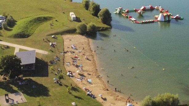 Vista aerea di una zona lacustre con spiaggia sabbiosa, un pendio erboso e un piccolo edificio.