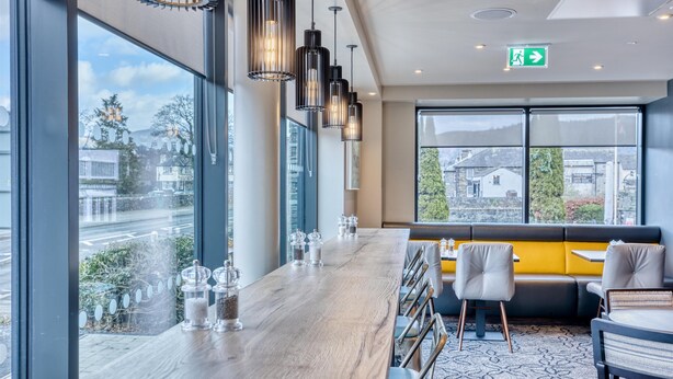 Un intérieur moderne avec un long bar en bois, un coin salon doté de coussins jaunes et de grandes fenêtres offrant une vue sur la ville.