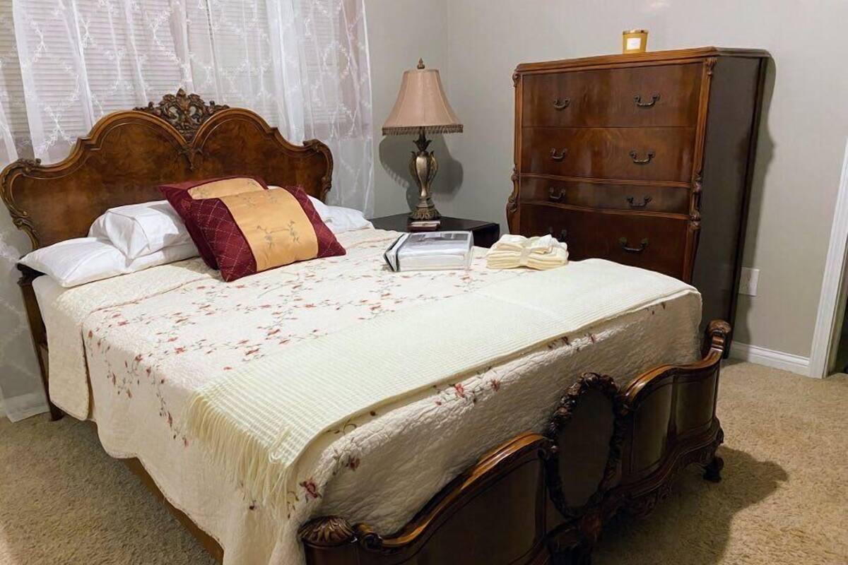 A neatly made bed with a floral bedspread and two pillows, a wooden dresser with multiple drawers, a bedside lamp, and a window with sheer curtains.