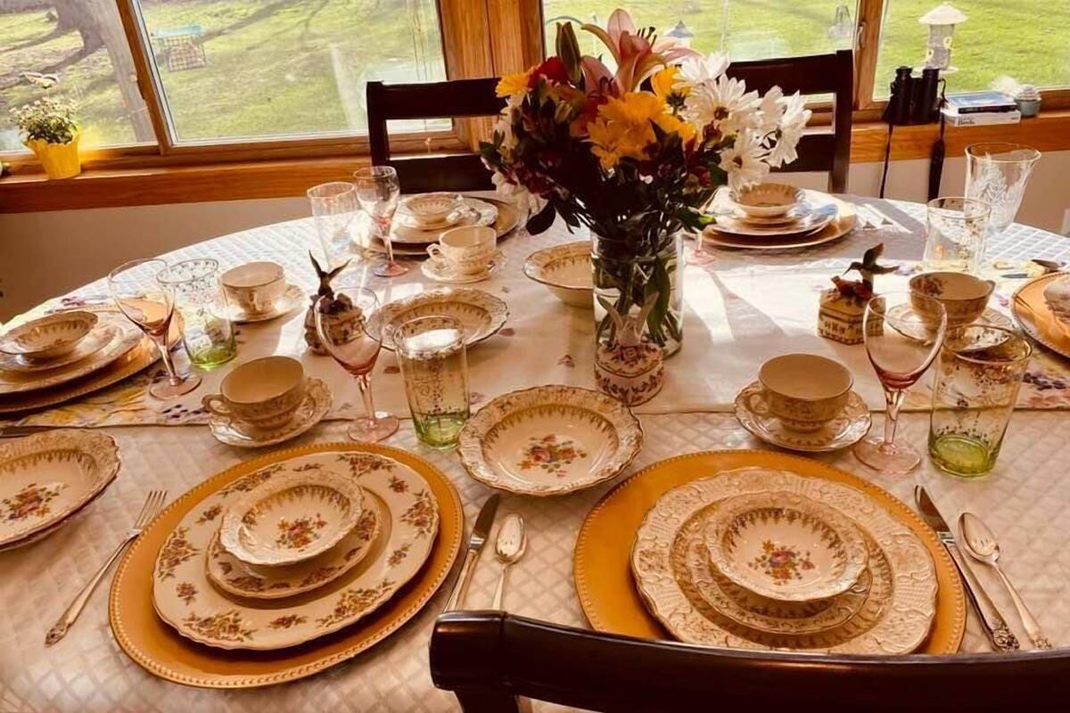 A round dining table set with gold-rimmed plates, glasses, and cutlery, centered with a vase of flowers.
