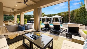 An outdoor lounge area with a sofa, chairs, a table with chess pieces, and umbrellas near a pool. - The Rose at Montage - Ultimate Estate (Indio)