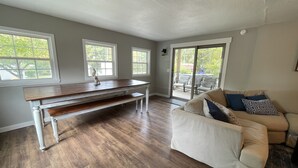 Un salón amplio con suelo de madera, un sofá blanco con cojines estampados, una mesa de centro de madera y un ventanal corredero que da a un patio exterior.